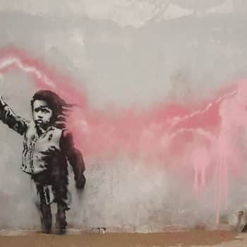 Migrant Child (2019), gesprühte Schablone von Banksy, in Venedig. Das Graffiti wurde während der Biennale von Venedig geschaffen und stellt ein Kind mit einer Schwimmweste und einer brennenden Fackel in der Hand dar. Es ist eine neue Anspielung auf das Leid der Migranten und Flüchtlinge, insbesondere derer, die am meisten leiden: der Kinder. Das Thema der Biennale von Venedig 2019 lautete „In interessanten Zeiten leben wollen“