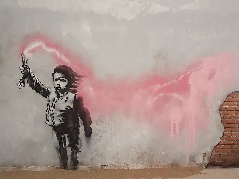 Migrant Child (2019), gesprühte Schablone von Banksy, in Venedig. Das Graffiti wurde während der Biennale von Venedig geschaffen und stellt ein Kind mit einer Schwimmweste und einer brennenden Fackel in der Hand dar. Es ist eine neue Anspielung auf das Leid der Migranten und Flüchtlinge, insbesondere derer, die am meisten leiden: der Kinder. Das Thema der Biennale von Venedig 2019 lautete „In interessanten Zeiten leben wollen“